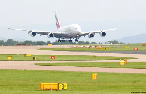 Emirates A380 landing This is a landscape photograph taken in late morning during the early autumn season at Heathrow Airport in the United Kingdom. The main subject of the image is an Emirates Airbus A380 aircraft, which is in the process of landing on the runway. The A380, a large jet used for international transport, is prominently featured with its landing gear deployed as it descends towards the tarmac. Various runway signs and markings can be seen across the airport grounds, and low, hazy hills rise in the background, indicating the outskirts of the airport. The image clearly showcases the scale of the Emirates aircraft and the infrastructure of Heathrow, emphasizing the airport’s role in global transport.
